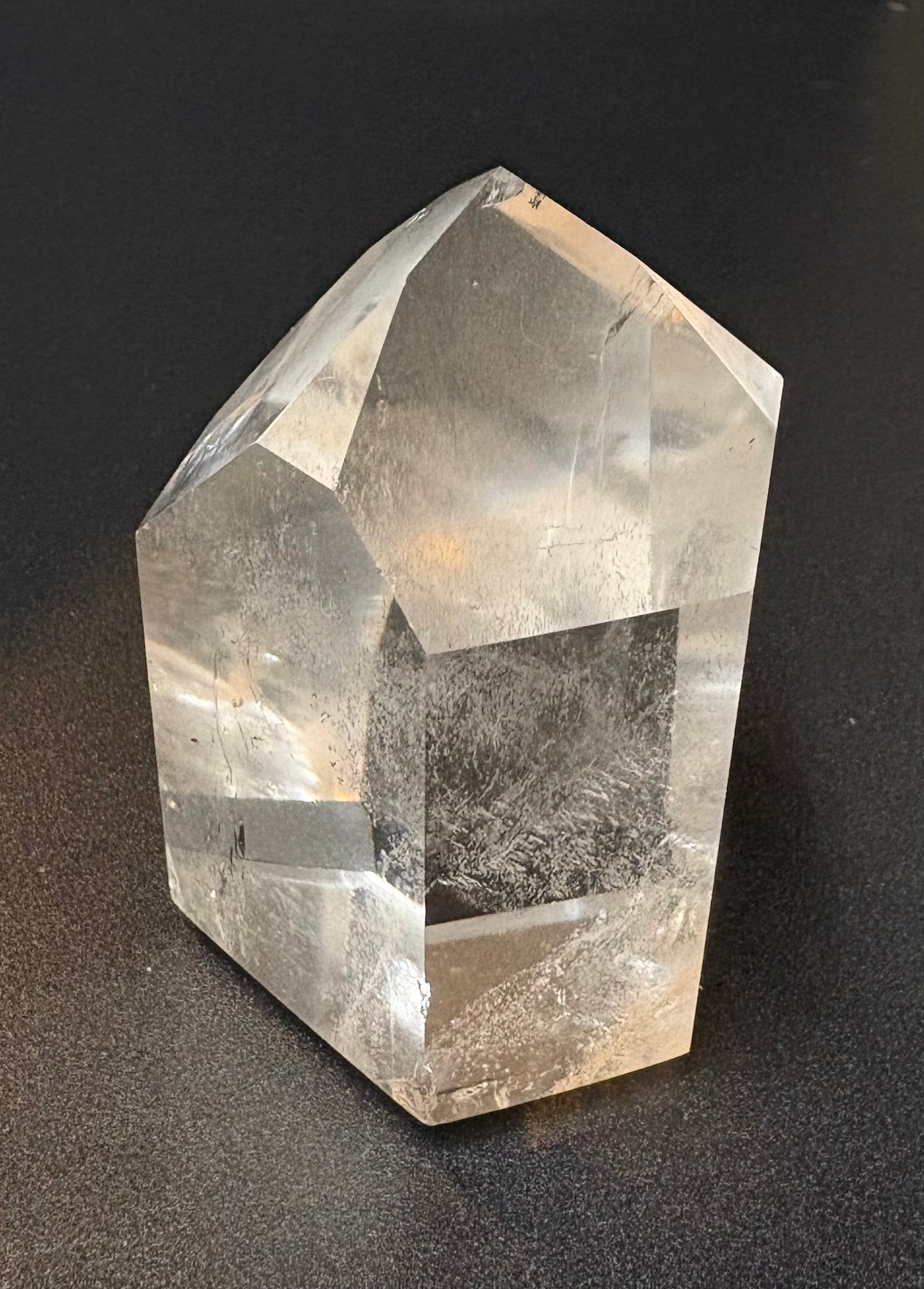 A clear faceted crystal point, likely quartz, standing upright on a dark textured surface.