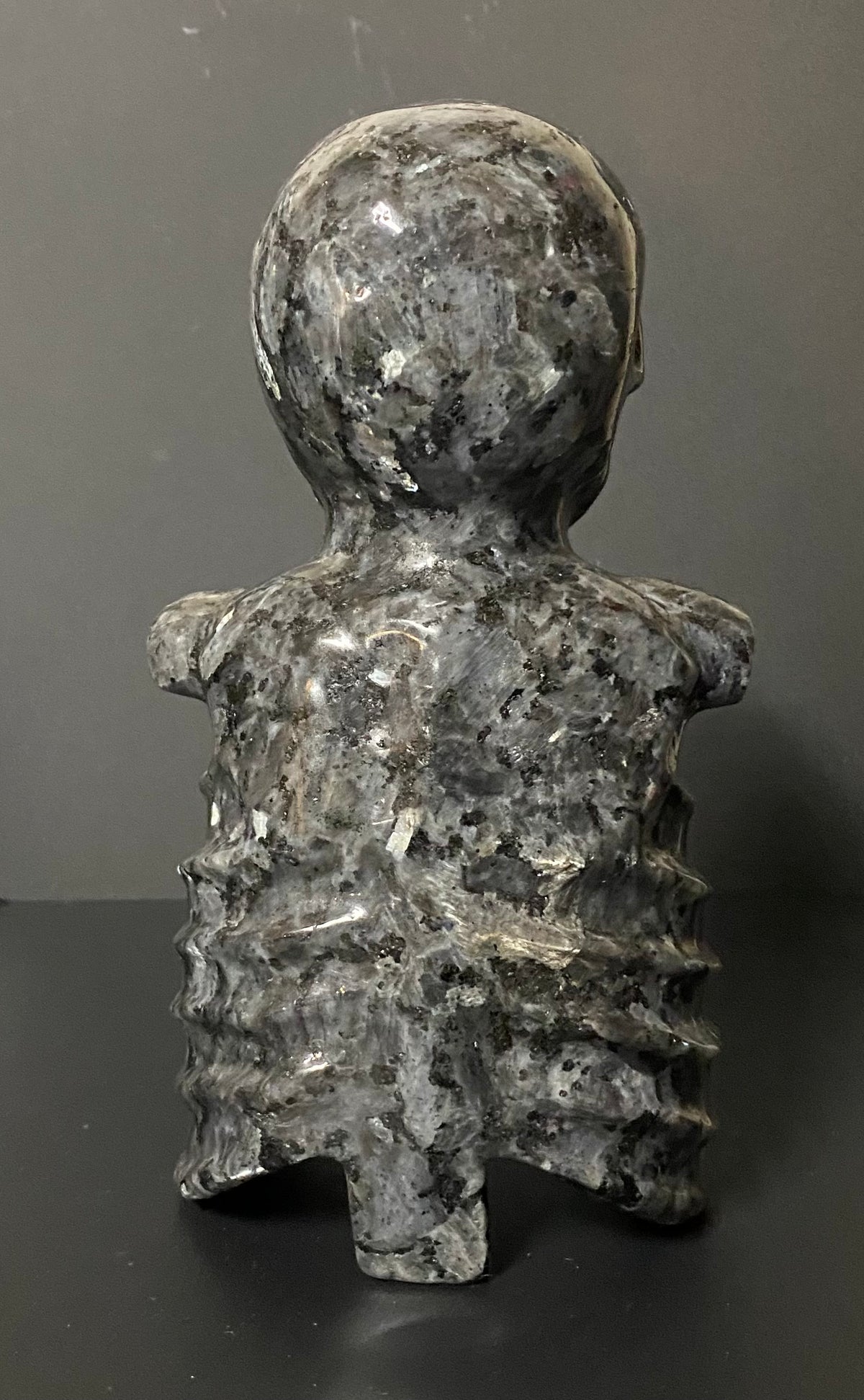 A Larvikite carved stone figurine of a skeleton back, made from polished gray Larvikite with natural speckled patterns, set against a dark gray background.