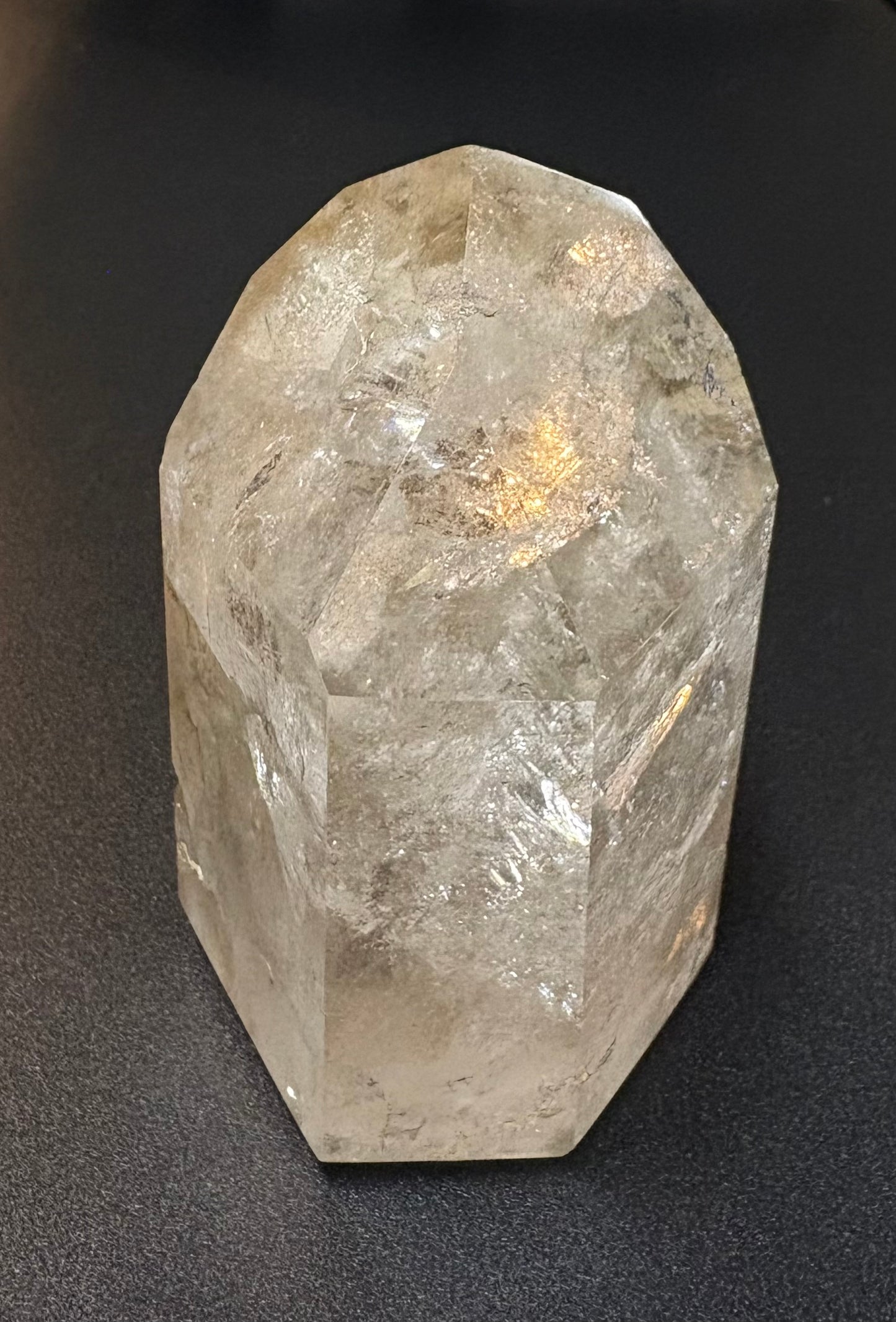 A raw, faceted clear quartz crystal tower standing upright on a dark, textured surface.