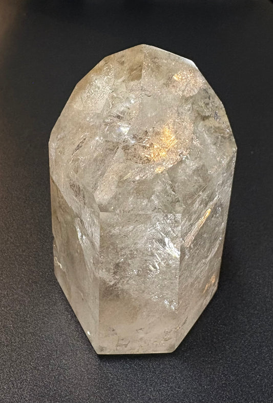 A raw, faceted clear quartz crystal tower standing upright on a dark, textured surface.