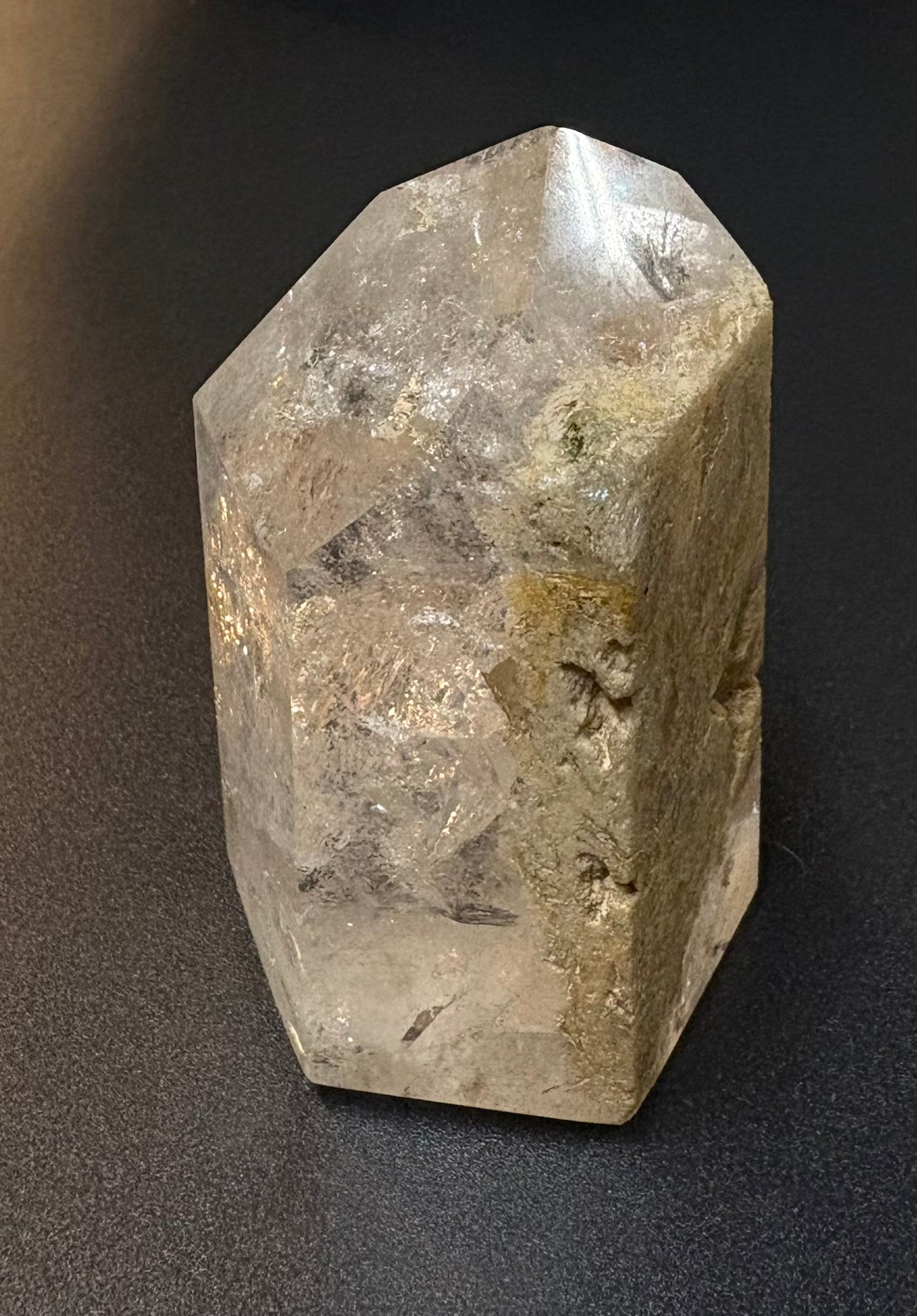 A natural clear quartz crystal point with internal inclusions and rough, textured edges, photographed on a dark gray background.