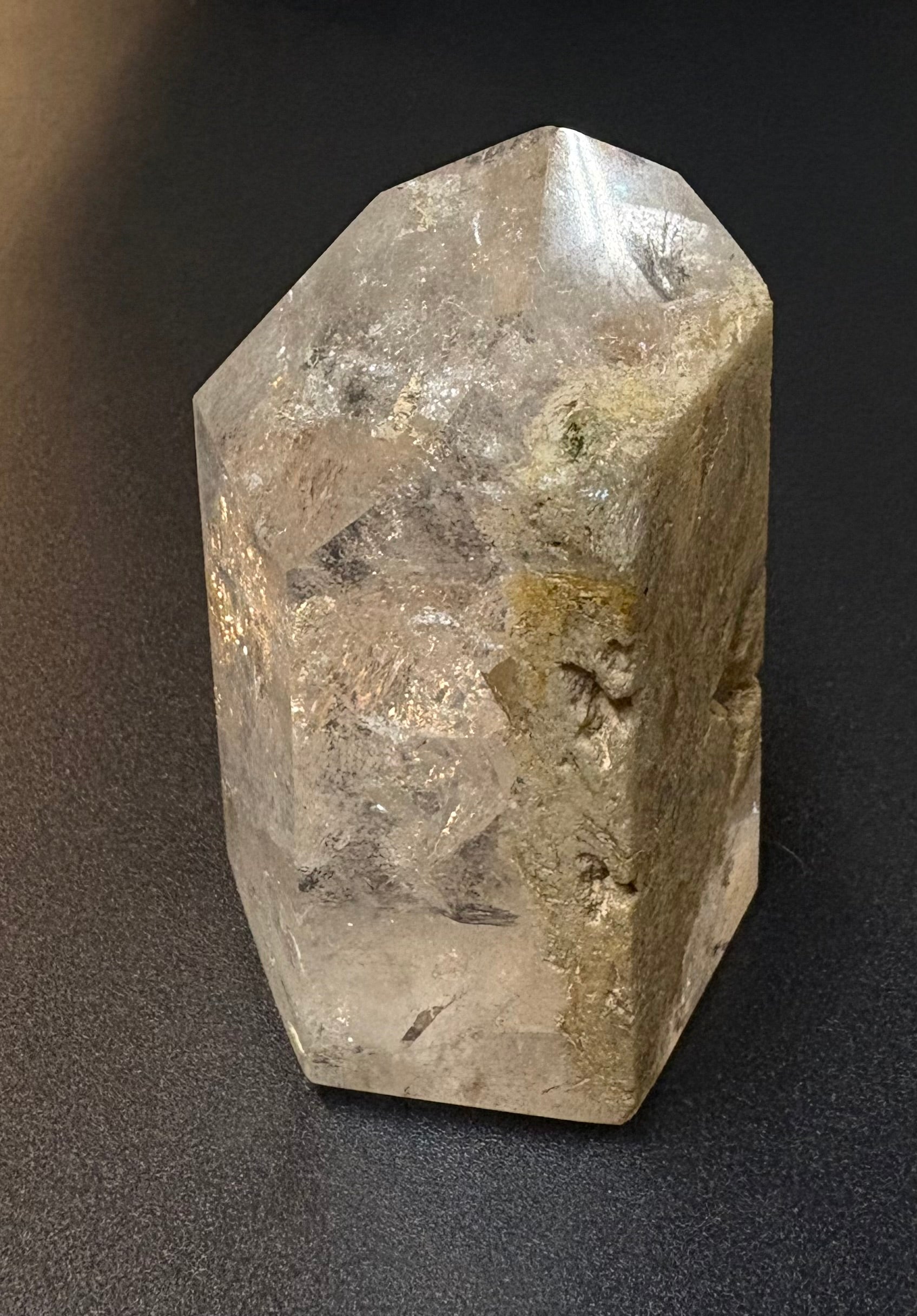 A natural clear quartz crystal point with internal inclusions and rough, textured edges, photographed on a dark gray background.