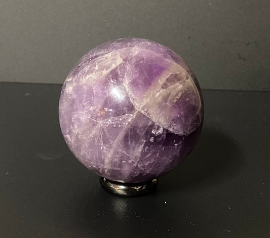 A polished purple amethyst crystal sphere resting on a small black metal stand against a dark gray background.