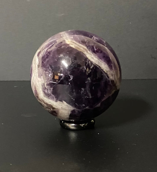 A polished purple amethyst crystal sphere with white veining, displayed on a small black stand against a dark gray background.