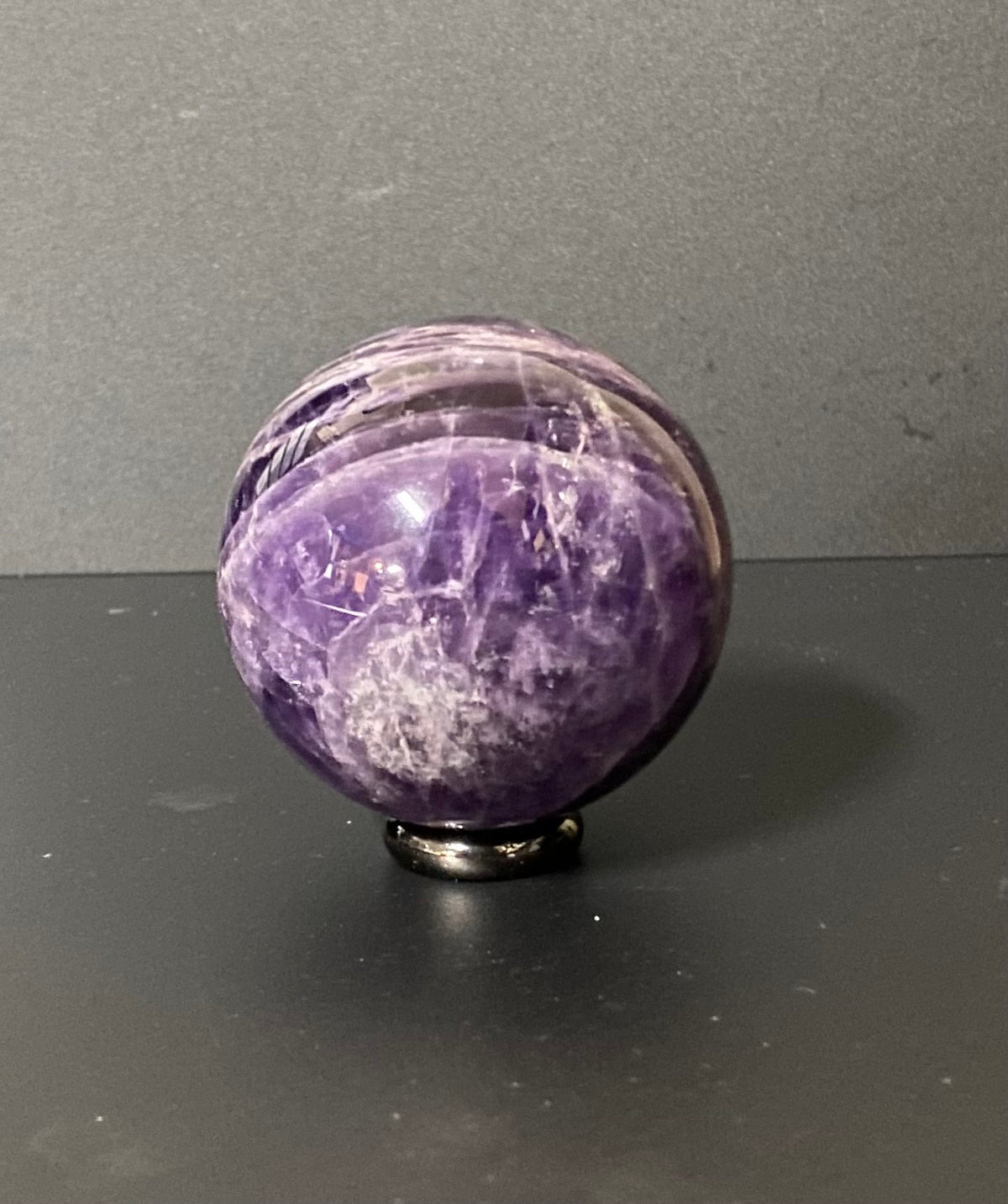 A polished purple amethyst crystal sphere resting on a small black metal stand against a dark gray background.