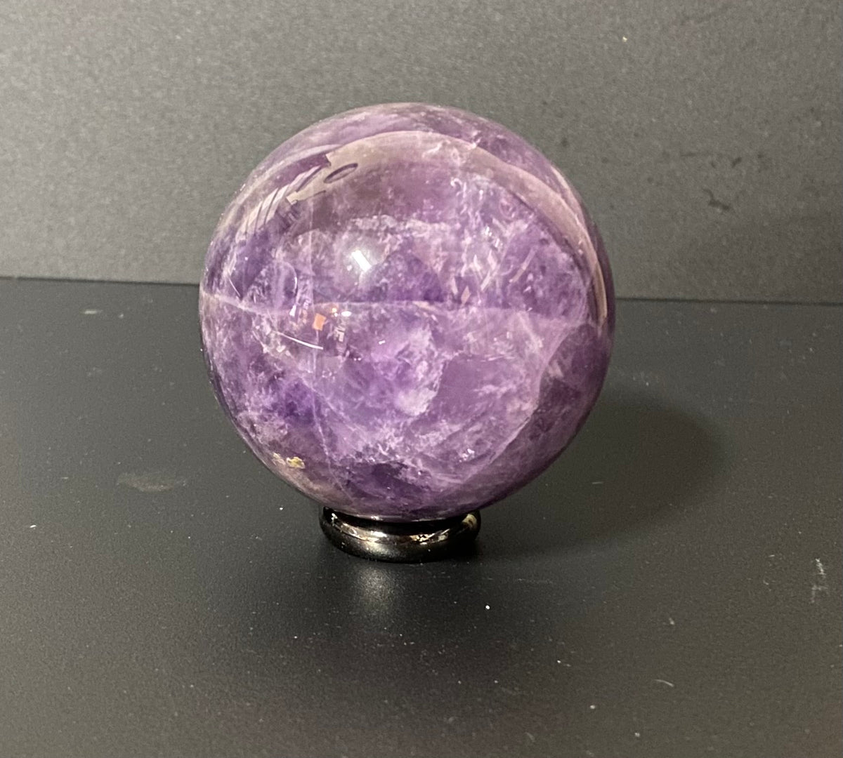 A polished purple amethyst crystal sphere resting on a small black metal stand against a dark gray background.