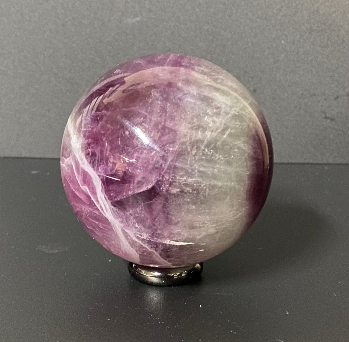 A polished fluorite crystal sphere with purple and white marbling, displayed on a small black stand against a dark gray background.