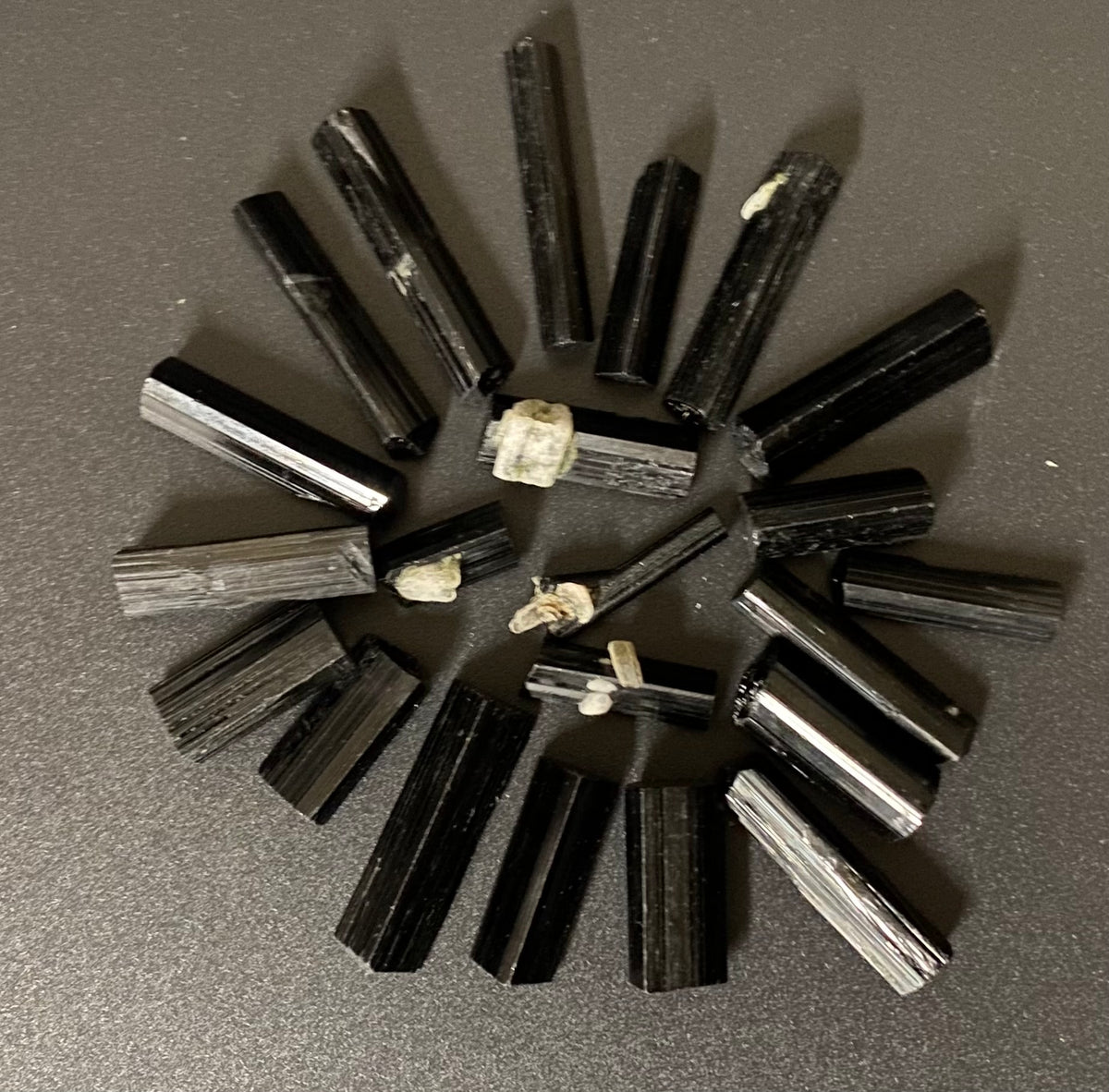 A circular arrangement of polished black tourmaline crystal rods, likely magnetite or similar ferrous mineral specimens, displayed on a dark gray textured surface.