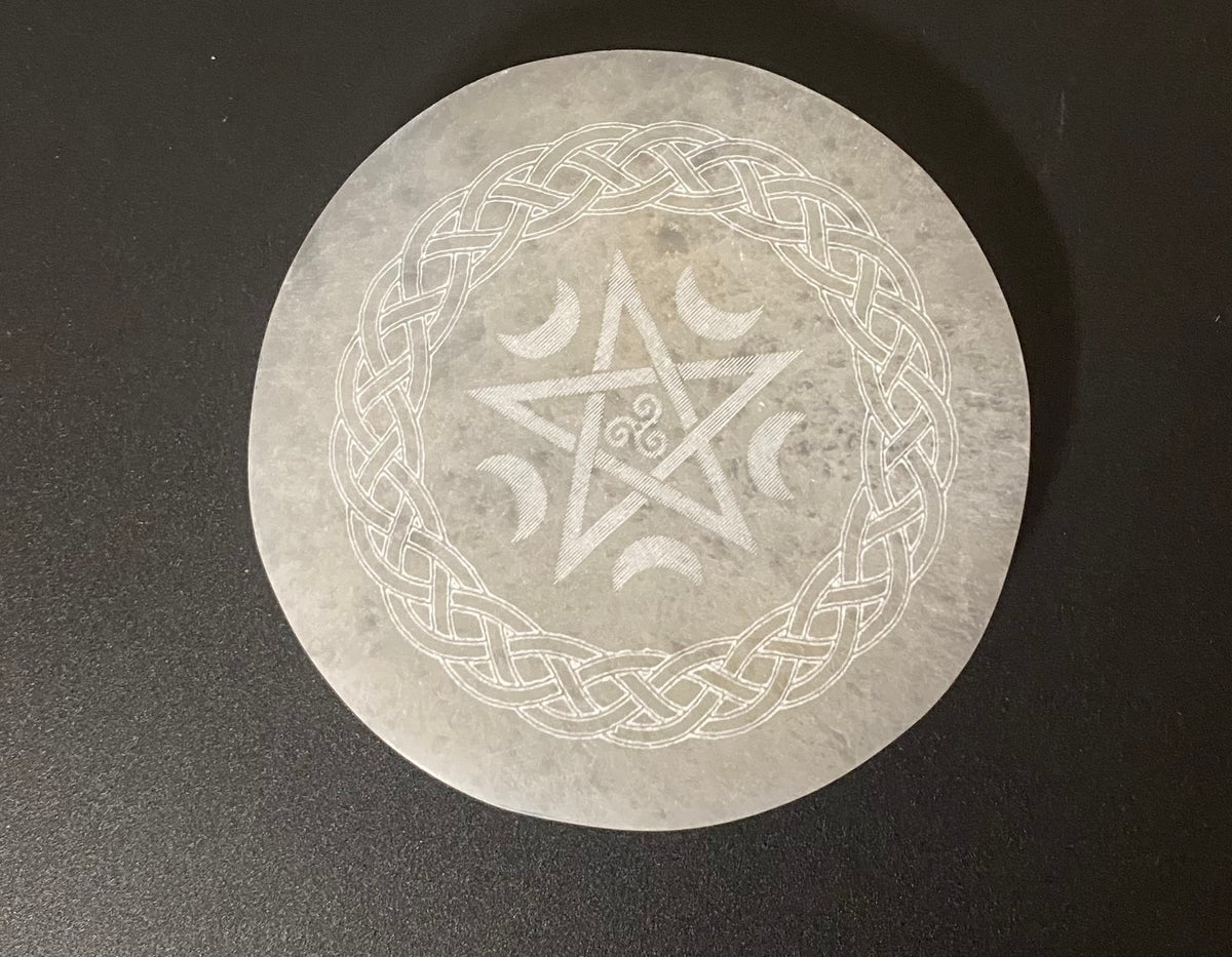 A circular stone selenite charging plate featuring a white engraved Celtic knot border, a central pentagram, and crescent moon symbols, displayed on a dark textured surface.