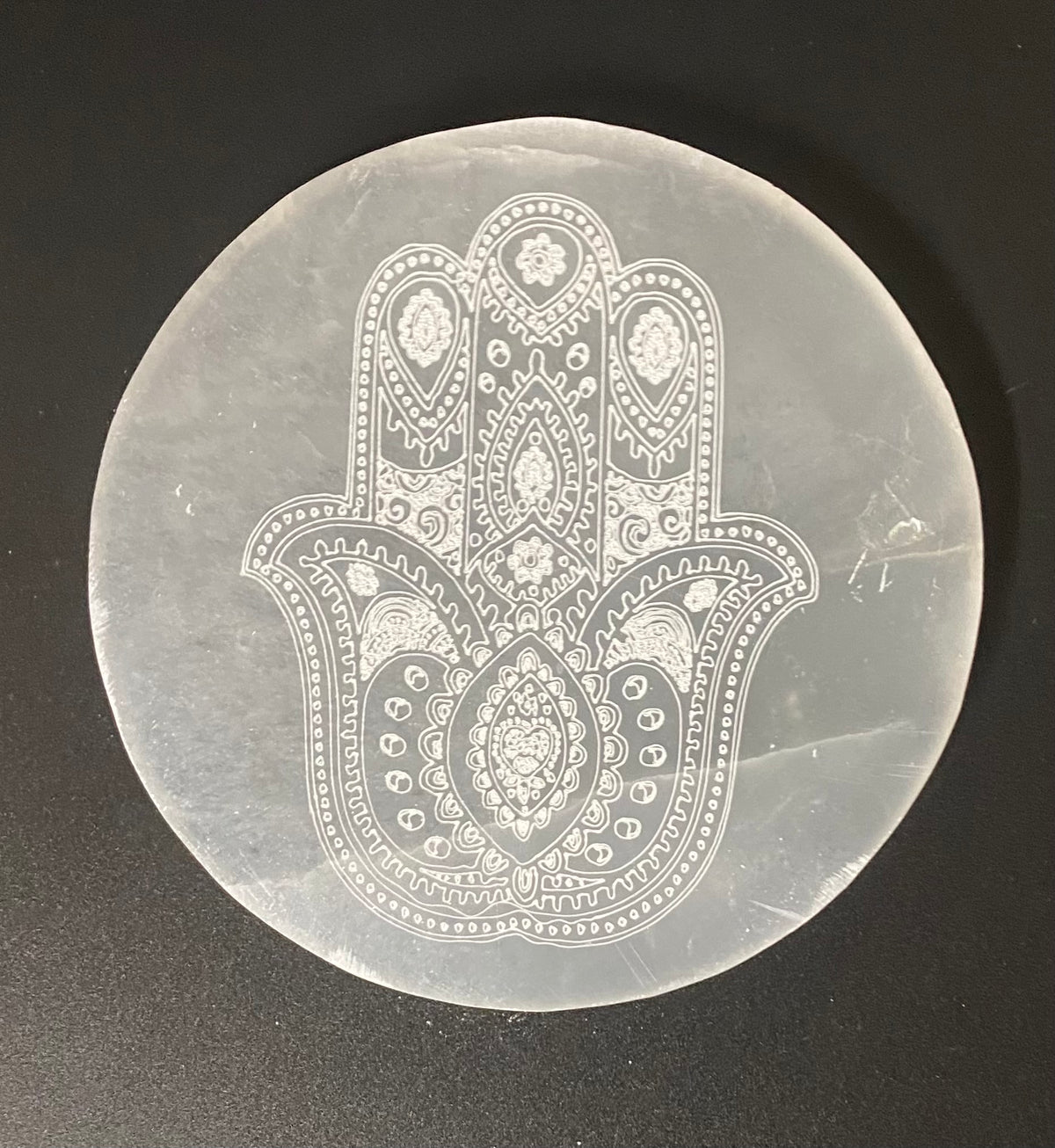 A circular selenite charging plate featuring a detailed white Hamsa hand design with intricate floral and geometric patterns, set against a dark gray background.