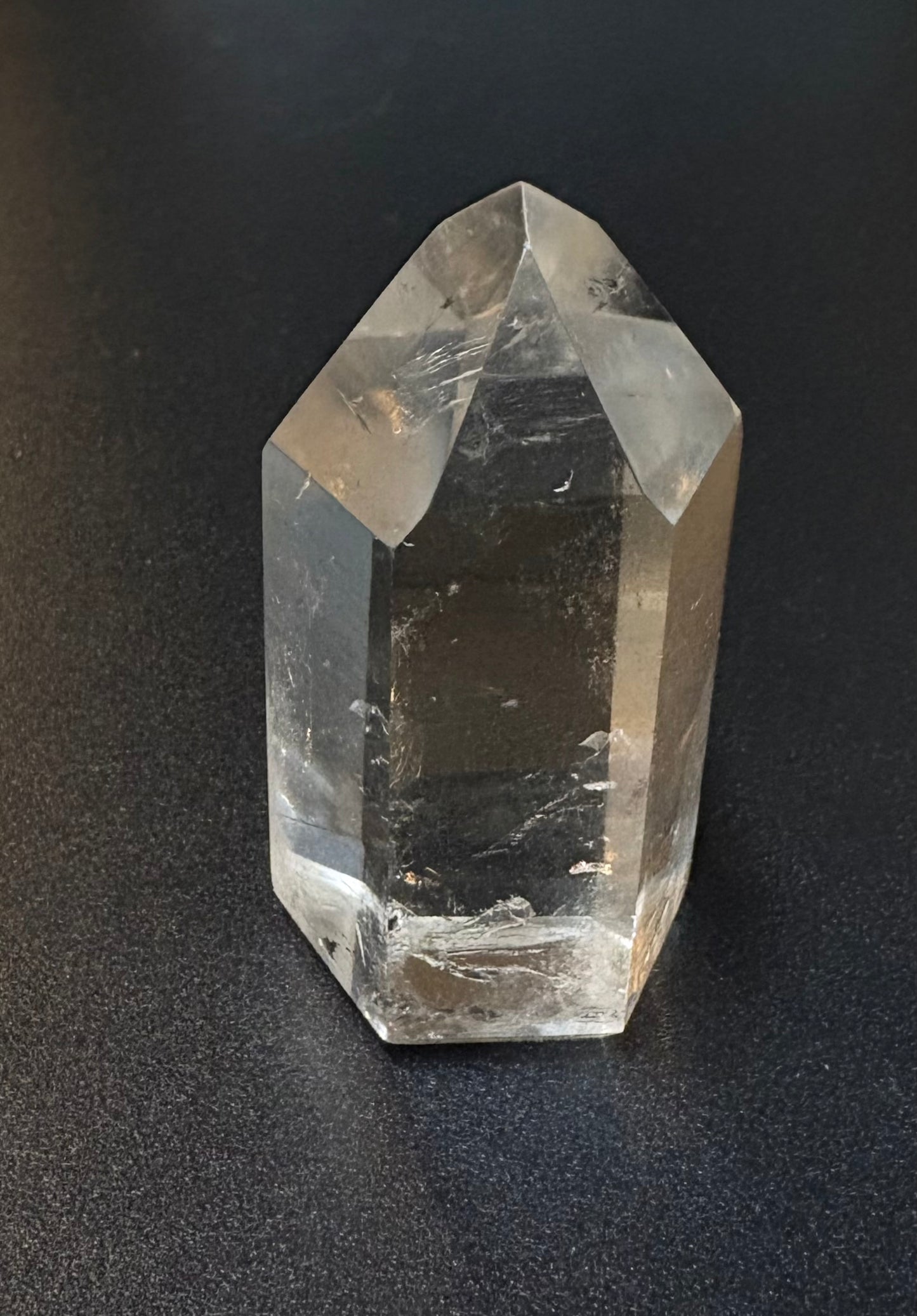 A natural clear quartz crystal point with sharp geometric facets, standing upright on a dark textured surface.