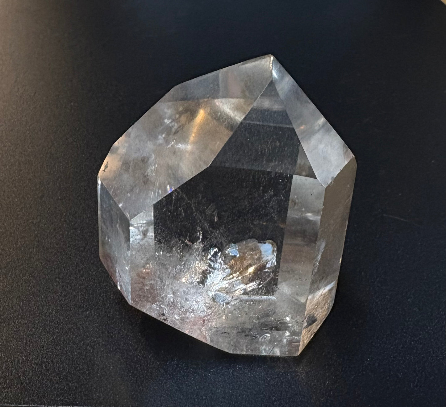 A faceted clear crystal stone, resembling a raw quartz point, displayed on a dark, textured background.