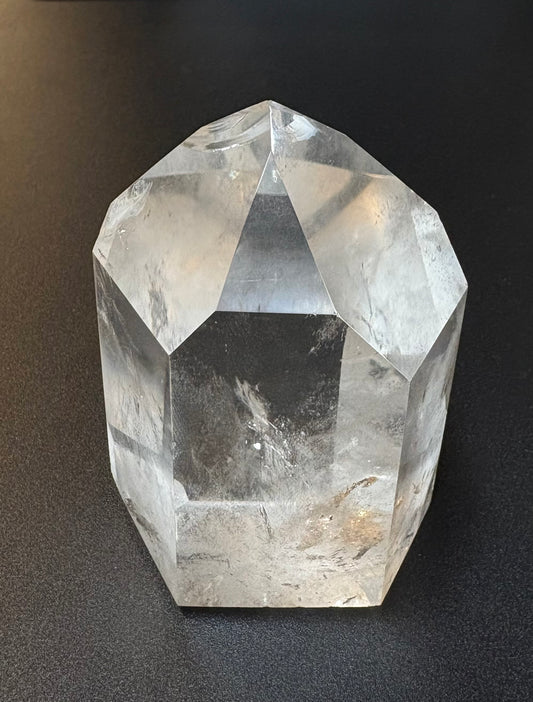A clear faceted crystal tower or point-shaped stone with geometric planes, photographed on a dark textured surface.