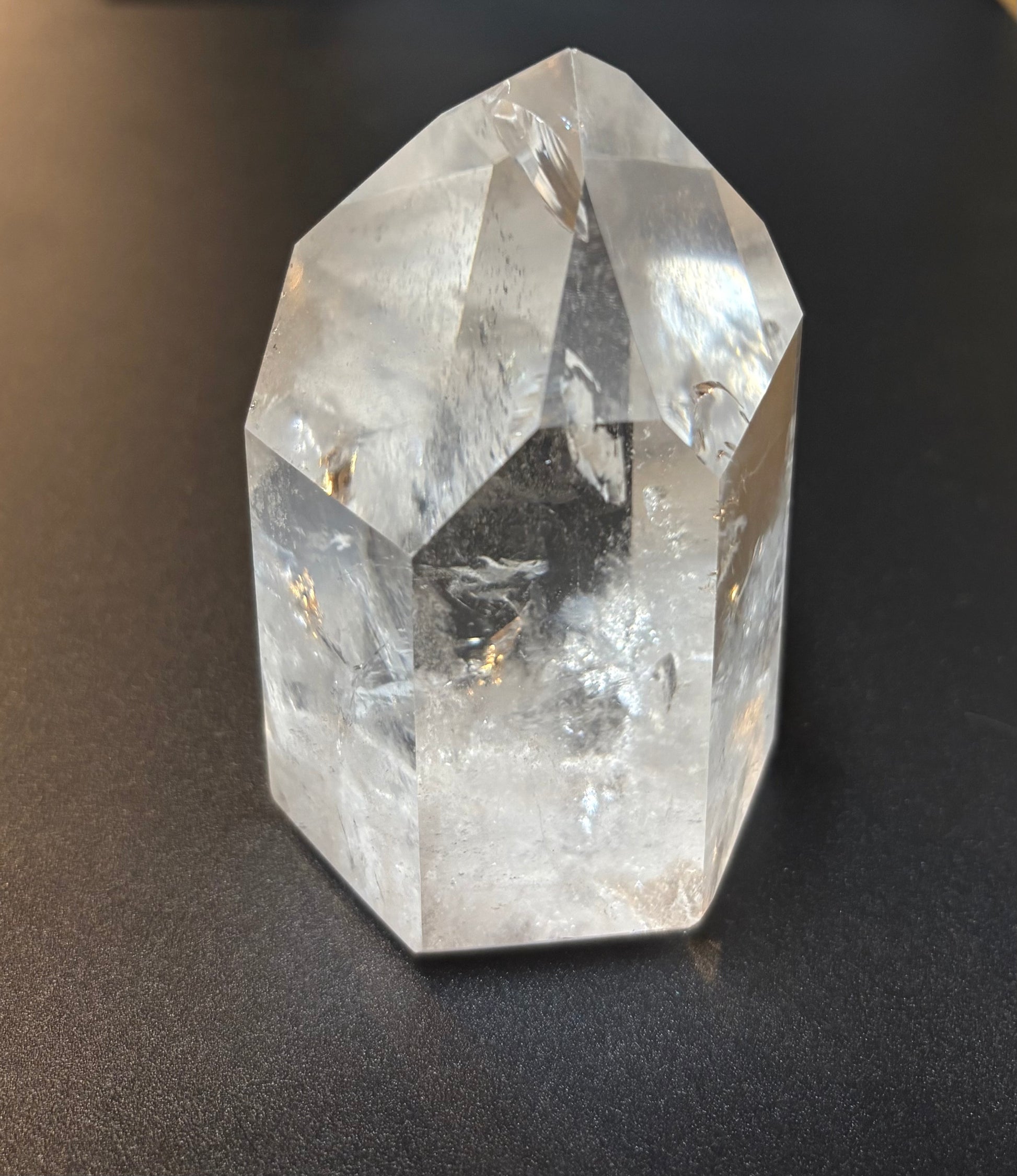A clear faceted crystal point, likely quartz, resting on a dark textured surface. The crystal is transparent with natural internal inclusions and reflections.