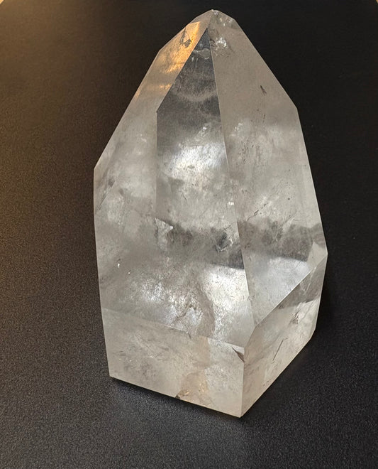 A natural clear quartz crystal point with internal inclusions, standing on a dark textured surface.