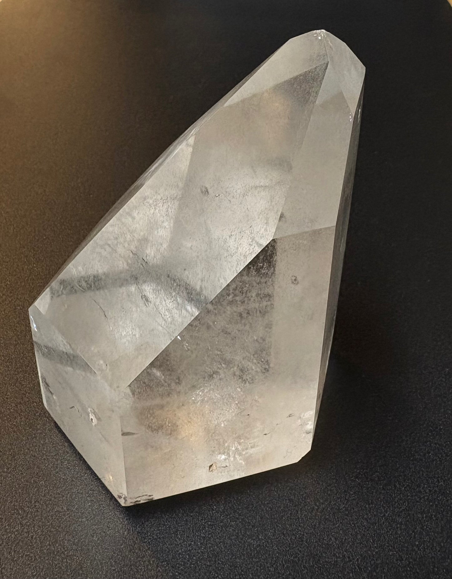 A clear faceted crystal point, likely quartz, resting on a dark textured surface.