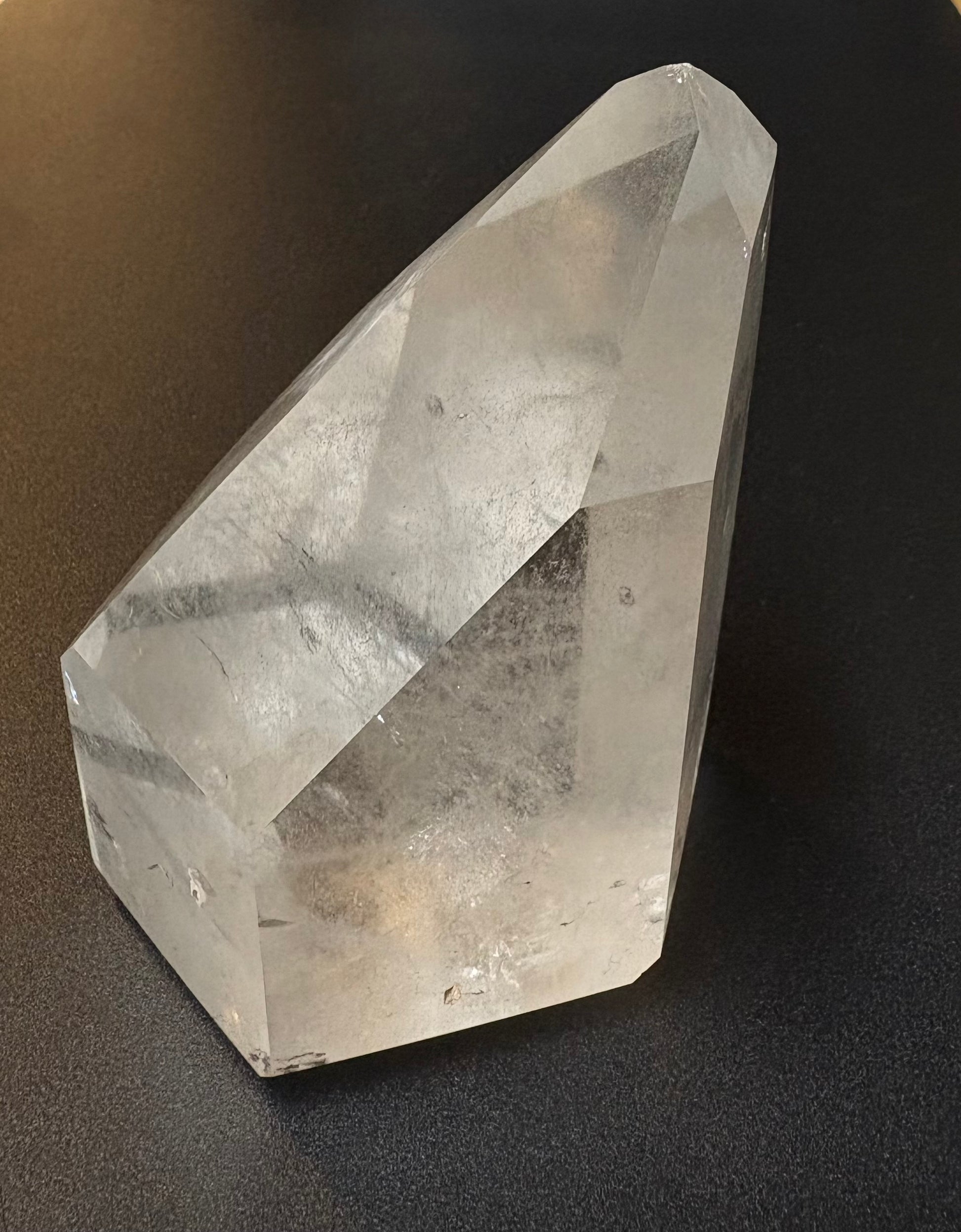 A clear faceted crystal point, likely quartz, resting on a dark textured surface.