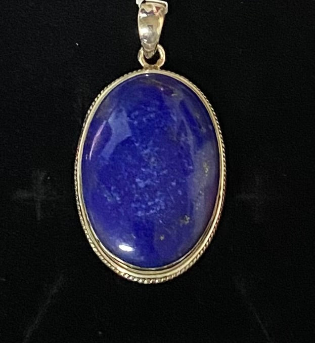 An oval pendant featuring a polished blue lapis lazuli stone set in a Sterling Silver bezel with a twisted rope border, shown against a black background.