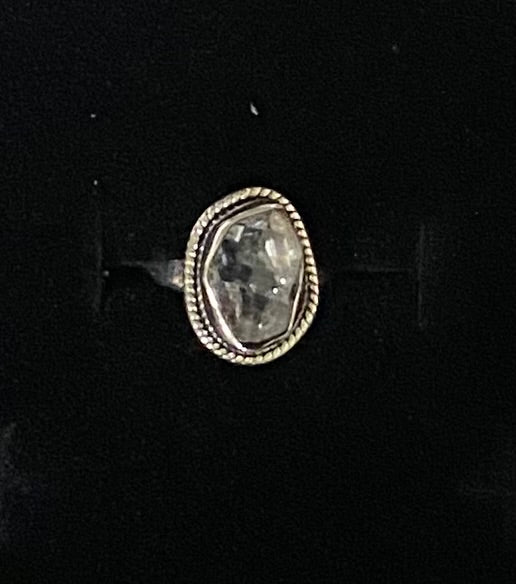 A silver-toned ring featuring a large, irregular, rough-cut clear gemstone in an oval bezel setting, displayed on a black background.