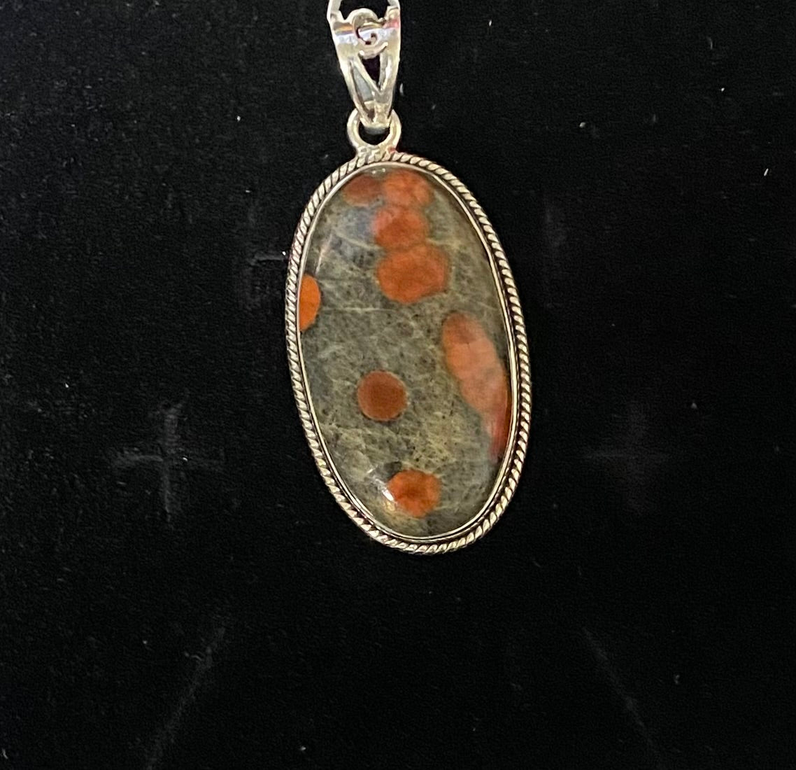An oval pendant featuring a natural stone with orange spots on a gray-green base, set in a silver-toned twisted bezel with a bail at the top, shown against a black background.