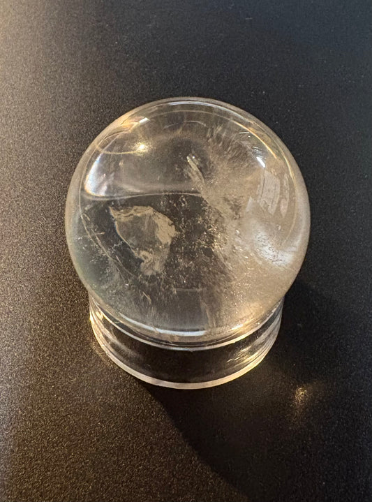 A quartz crystal ball with a circular base, photographed on a dark black surface. The ball appears transparent with some internal cloudy or crystalline texture.