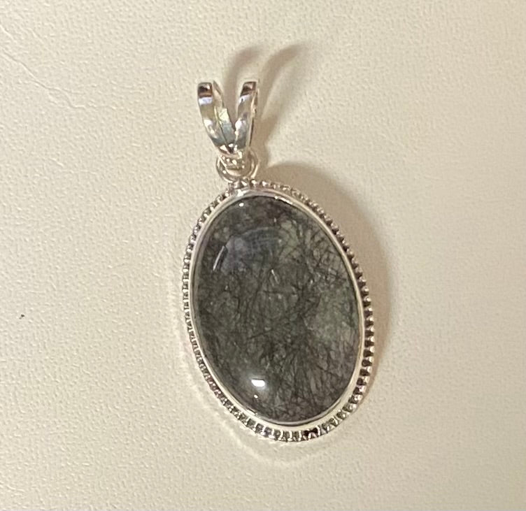 An oval pendant featuring a dark gray to black fibrous stone, set in a silver bezel with a beaded border and a silver bail, shown on a textured off-white background.