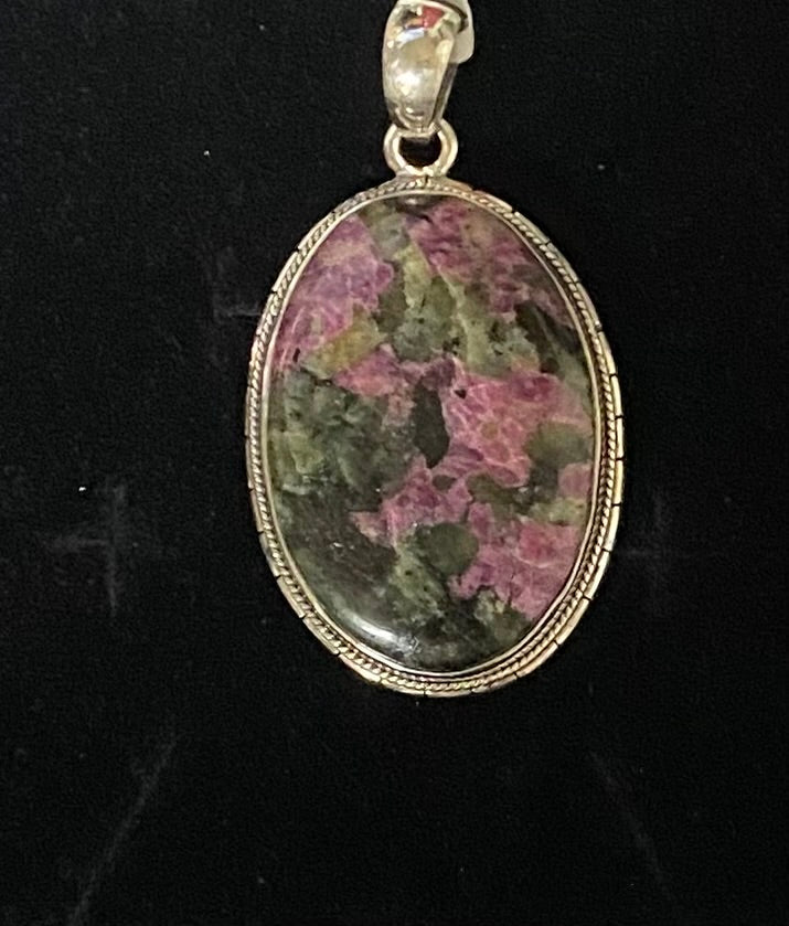 An oval pendant featuring a pink and green mottled stone, set in a silver-toned metal bezel with a bail at the top, shown against a black background.