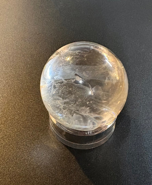 A clear quartz crystal ball with natural inclusions, resting on a clear acrylic stand against a dark gray background.