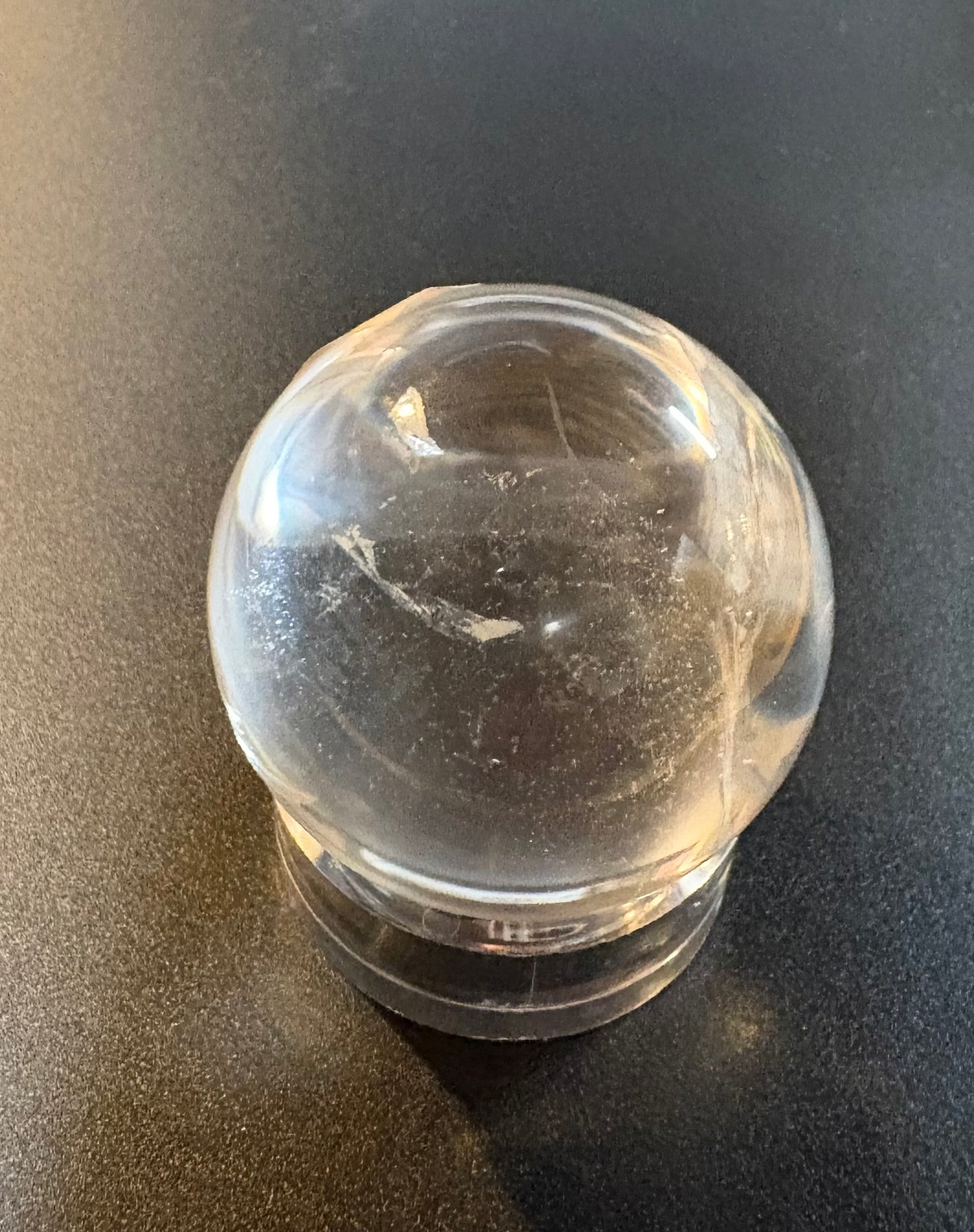 A clear quartz crystal ball with natural inclusions, resting on a clear acrylic stand against a dark gray background.