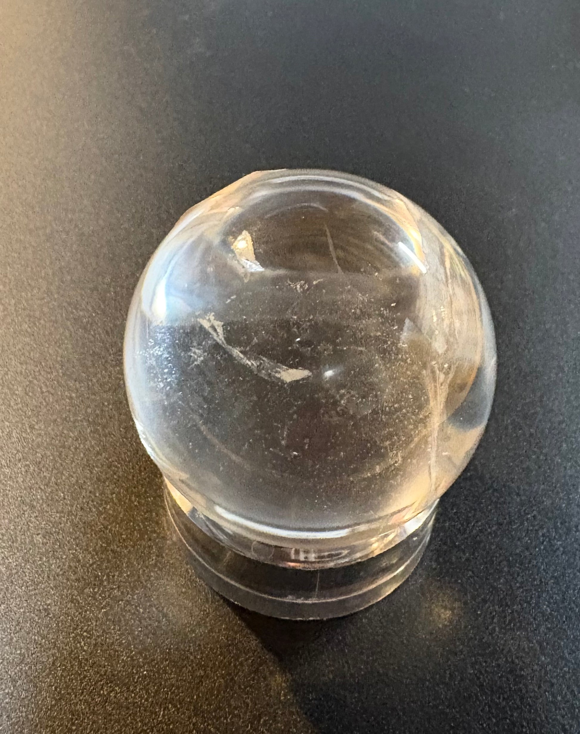 A clear quartz crystal ball with natural inclusions, resting on a clear acrylic stand against a dark gray background.