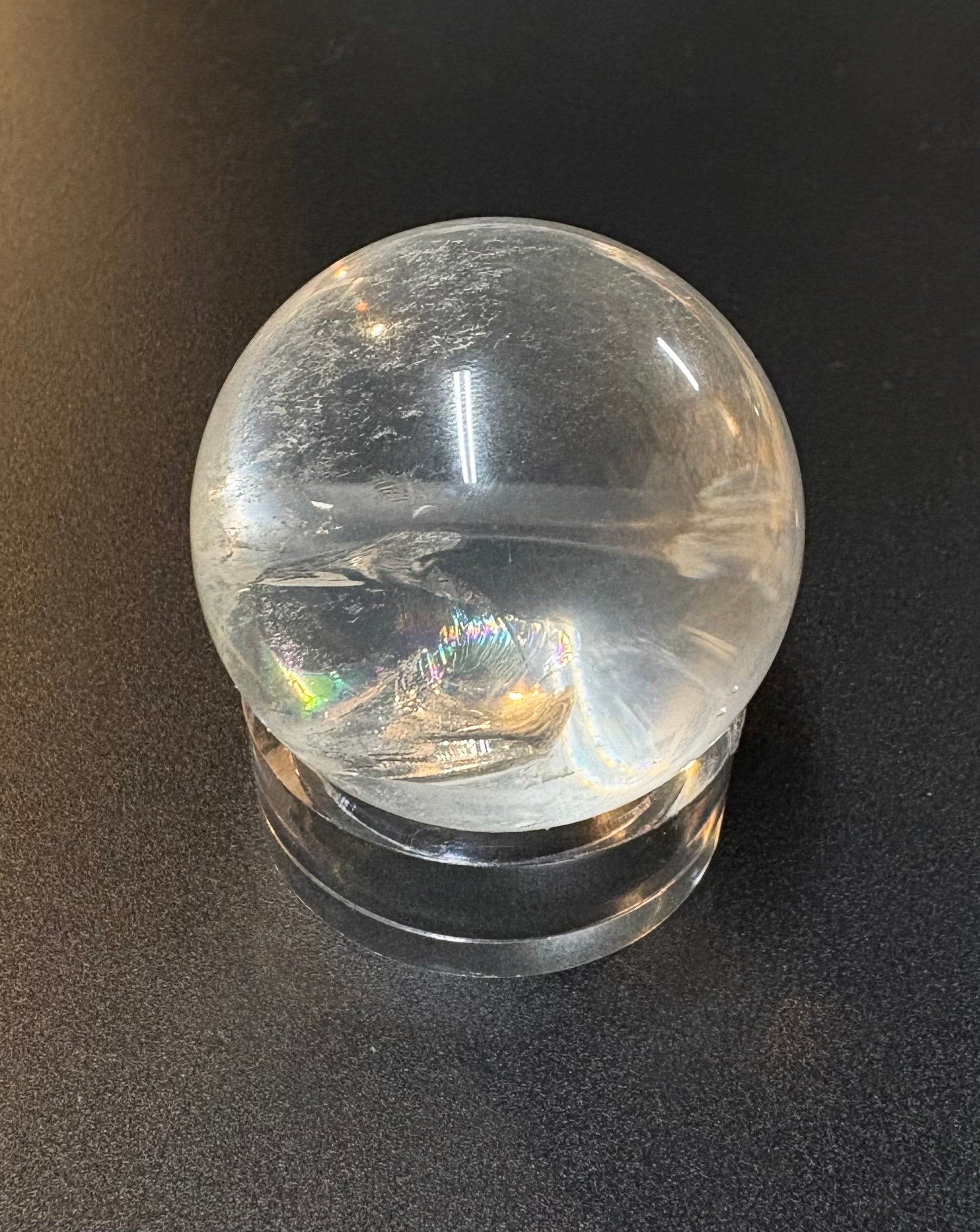 A clear quartz crystal ball with natural inclusions resting on a matching clear acrylic stand, photographed against a dark textured background.