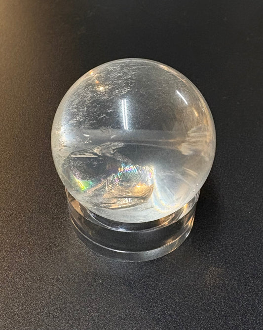 A clear quartz crystal ball with natural inclusions resting on a matching clear acrylic stand, photographed against a dark textured background.