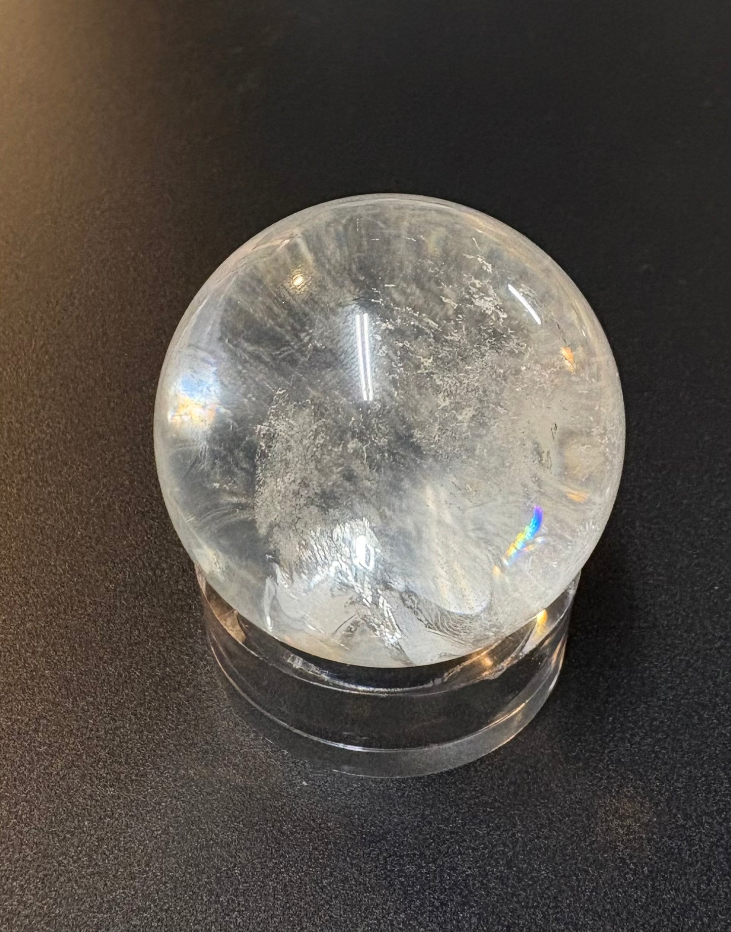 A clear quartz crystal ball with natural inclusions resting on a matching clear acrylic stand, photographed against a dark textured background.
