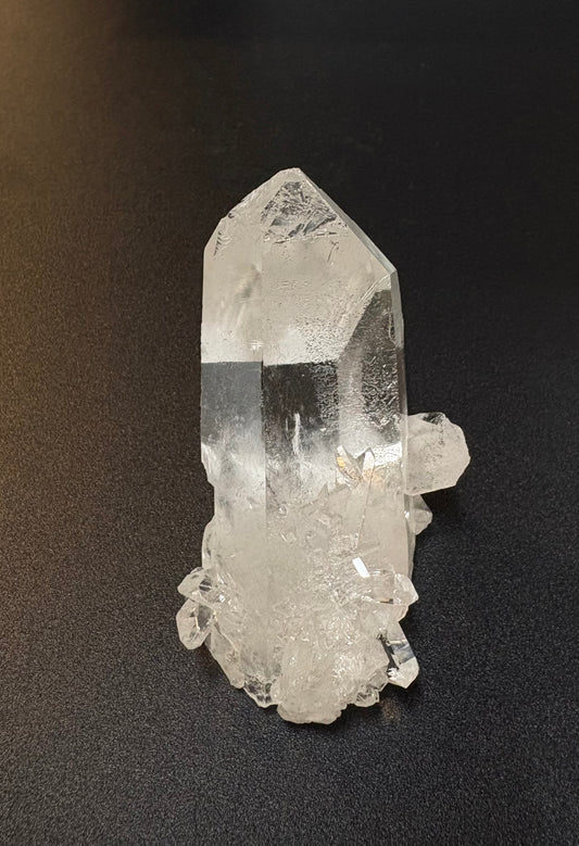 A natural clear quartz crystal point with rough, textured matrix base, photographed on a dark textured surface.