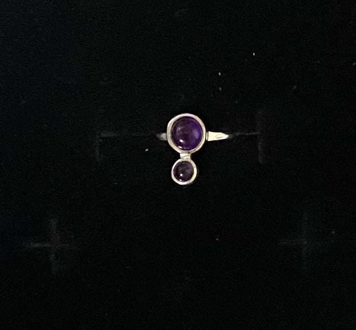 A silver ring featuring a large oval purple gemstone and a smaller round purple gemstone below it, displayed on a black background.