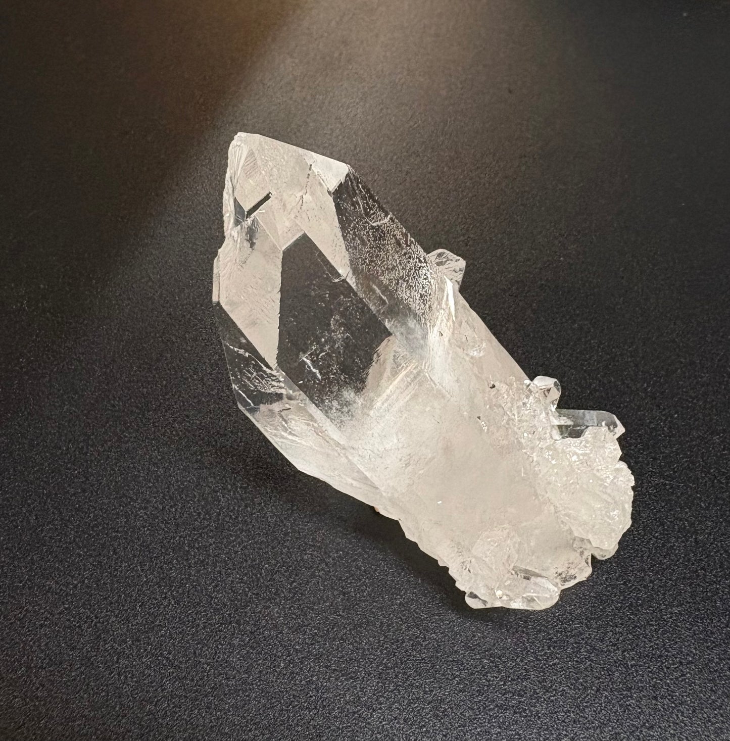 A raw, natural clear quartz crystal cluster with rough, unpolished edges, displayed on a dark textured background.