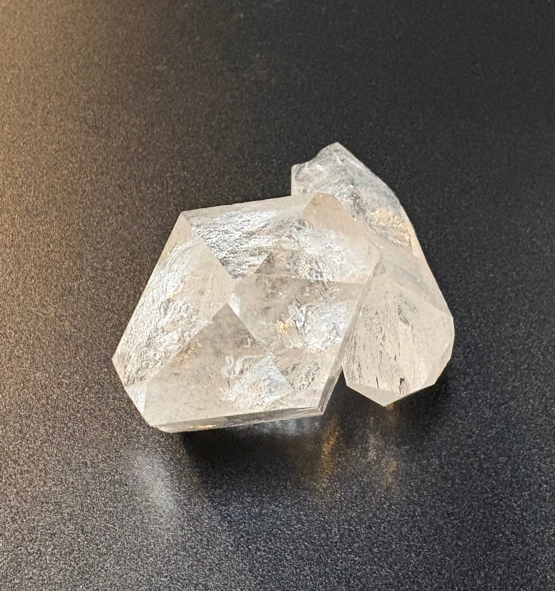 Clear faceted crystal stone with natural rough edges, resting on a dark textured surface.