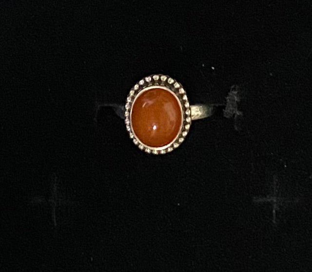 A vintage-style oval ring featuring a warm orange cabochon stone surrounded by a silver-tone beaded halo, set against a black background.