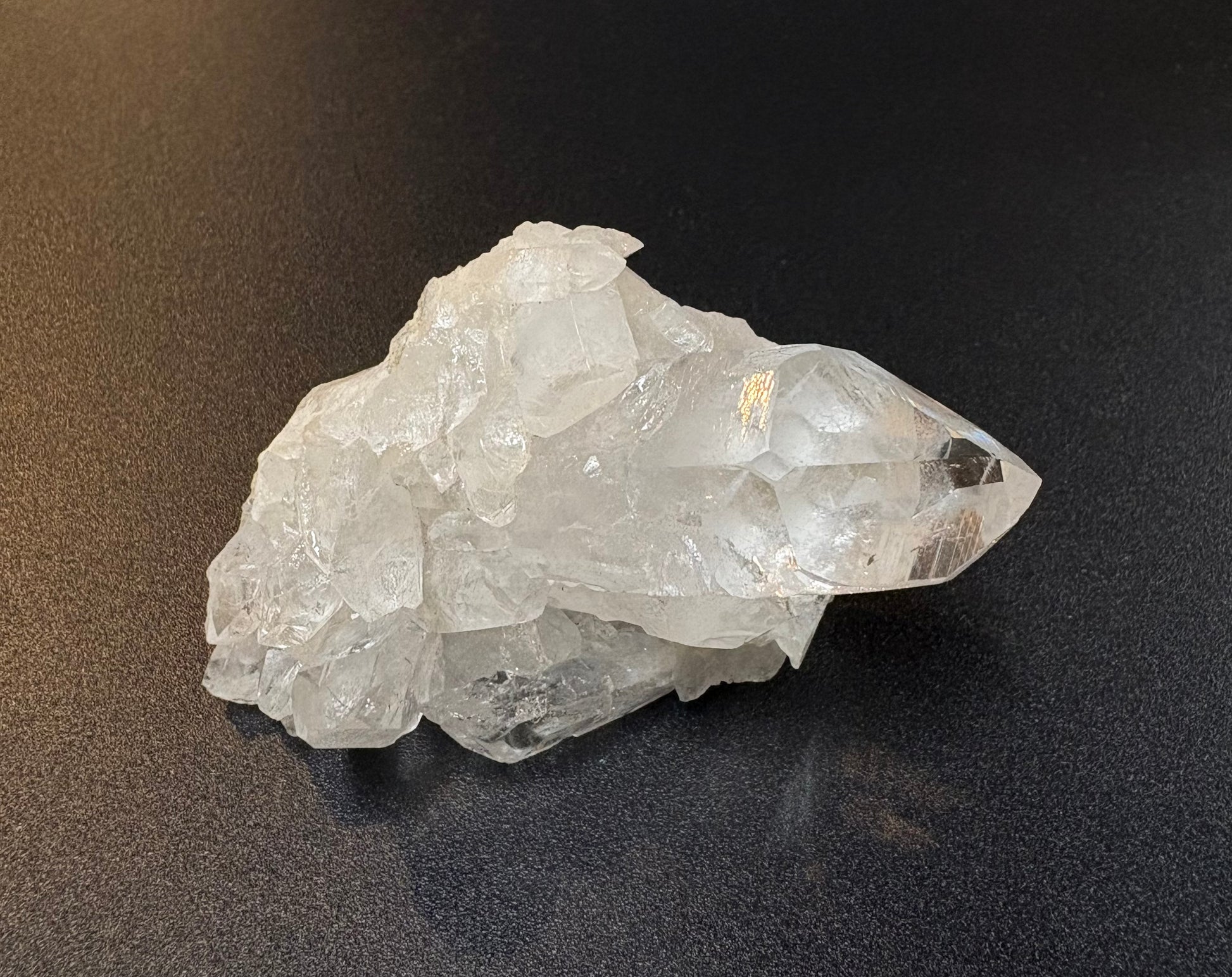 A raw, rough crystal cluster with a prominent pointed clear quartz point, resting on a dark, textured surface.