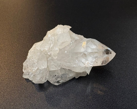 A raw, rough crystal cluster with a prominent pointed clear quartz point, resting on a dark, textured surface.