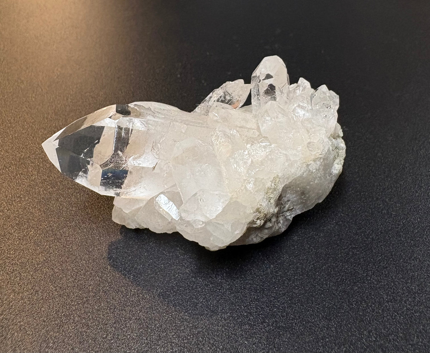 A clear quartz crystal cluster with a prominent pointed crystal point, resting on a dark textured surface.