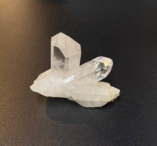 A cluster of natural clear quartz crystals on an unpolished base, photographed on a dark textured surface.