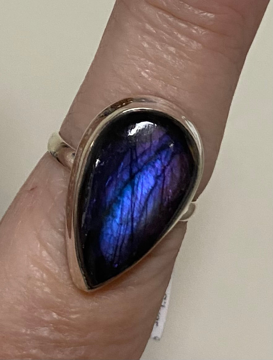 A silver ring featuring a large teardrop-shaped labradorite stone with a vibrant blue and purple iridescent shimmer, shown worn on a finger.