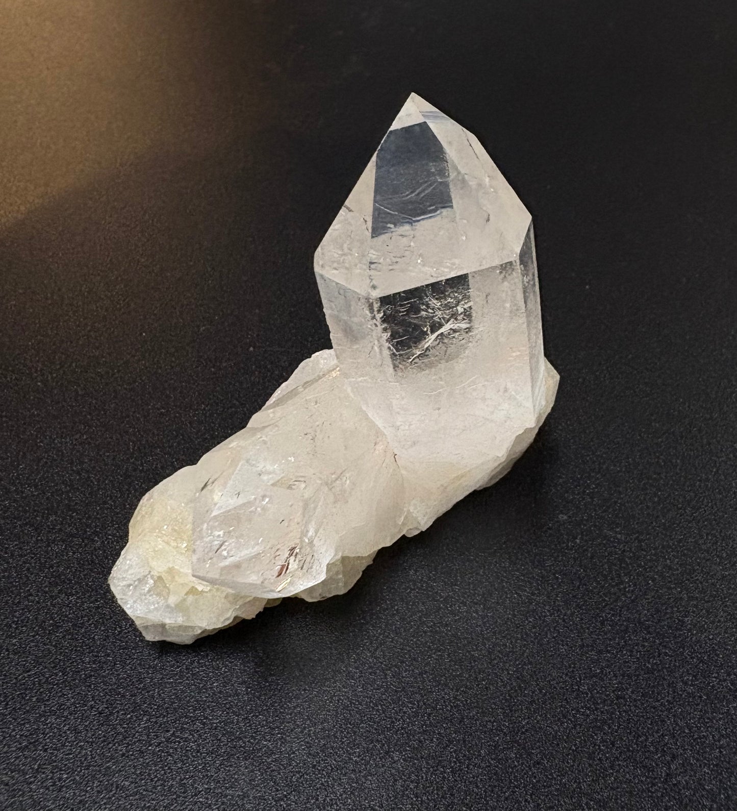 A natural clear quartz crystal cluster with a tall pointed secondary crystal on top of a rough white quartz base, photographed on a dark textured surface.