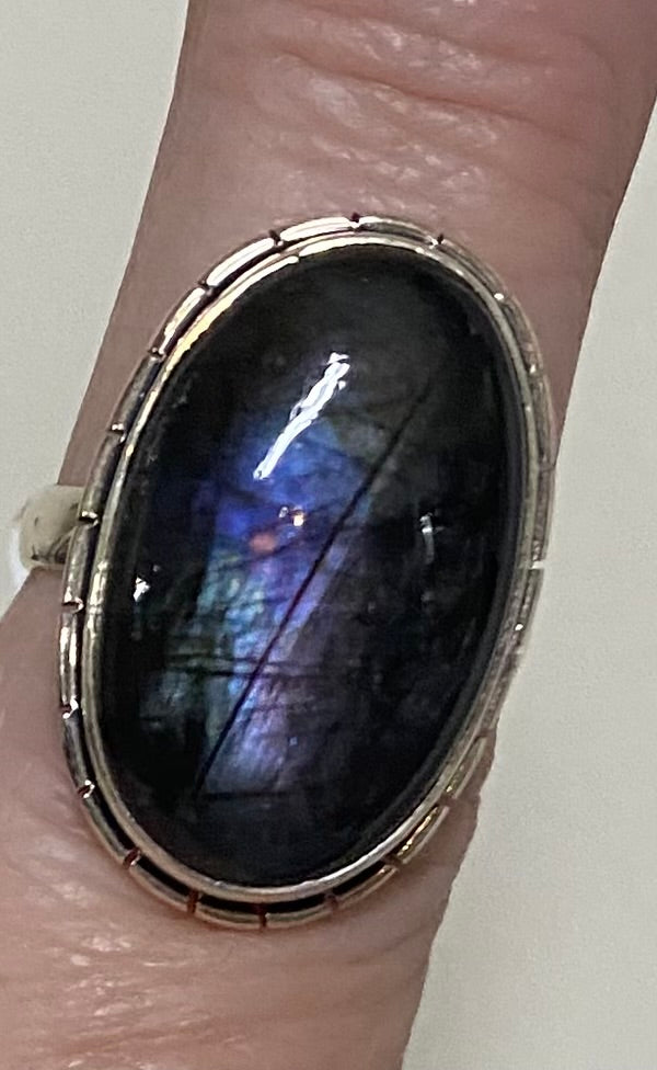 A close-up of a silver-toned ring with a large oval dark labradorite stone showing blue and purple iridescent flashes, worn on a finger.