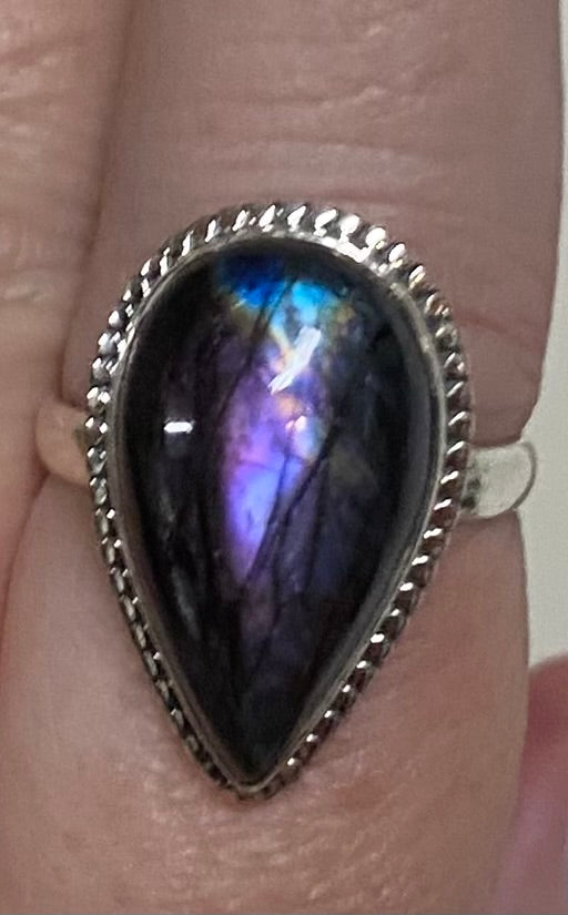 A close-up of a silver teardrop ring with a dark iridescent labradorite stone, worn on a finger.