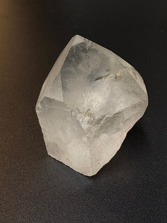 A raw, unpolished clear quartz crystal with natural geometric facets, resting on a dark textured surface.
