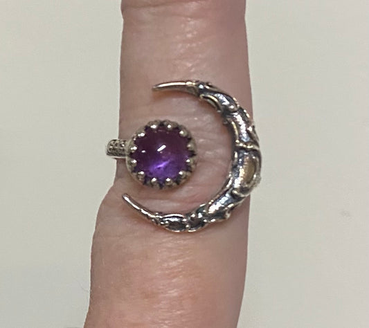 A silver-toned ring shaped like a crescent moon wrapping around a finger, featuring a round purple amethyst gemstone in a textured bezel setting.