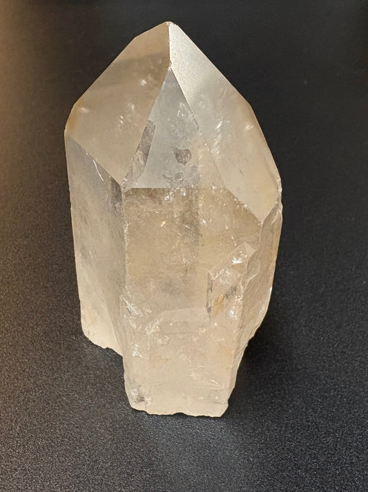 A raw, unpolished clear quartz crystal point resting on a dark gray textured surface.