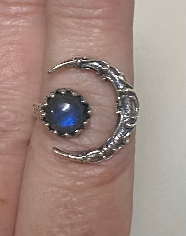 A close-up of a silver crescent moon ring worn on a finger, featuring a central oval blue-gray stone with a halo of small clear stones.